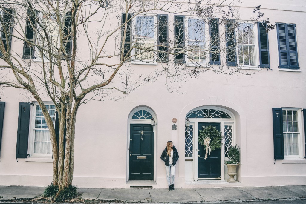 a young woman posing in front of a pastel pink house with black window shutters